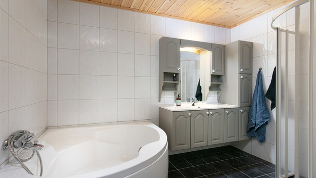 Badet har flislagt gulv med varmekabler, hjørnebadekar, dusjhjørne og grå baderomsinnredning.