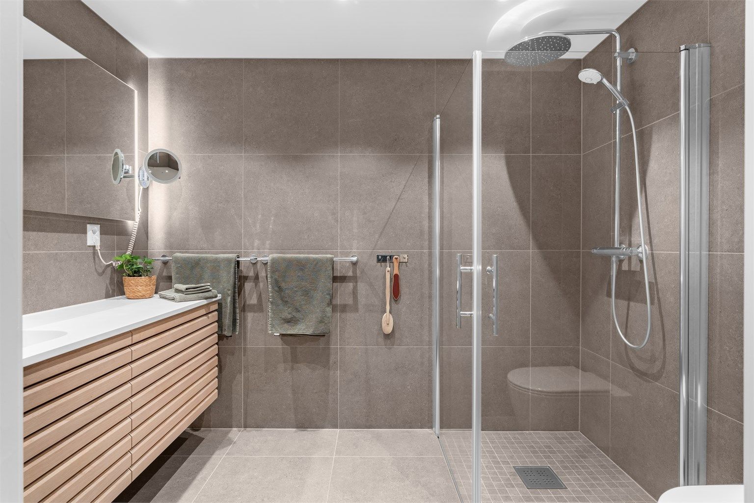 Badet har dusjnisje med glassdører og ett-greps dusjarmatur. Vegghengt toalett med innebygget sisterne.