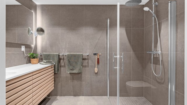 Badet har dusjnisje med glassdører og ett-greps dusjarmatur. Vegghengt toalett med innebygget sisterne.