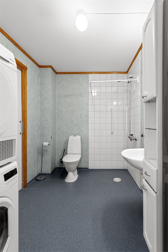 Videre er badet utstyrt med gulvstående toalett (ca. 2021-2022) og dusj i hjørne med veggmontert dusjarmatur samt opplegg for dusjforheng.