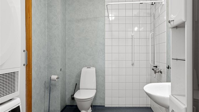 Videre er badet utstyrt med gulvstående toalett (ca. 2021-2022) og dusj i hjørne med veggmontert dusjarmatur samt opplegg for dusjforheng.