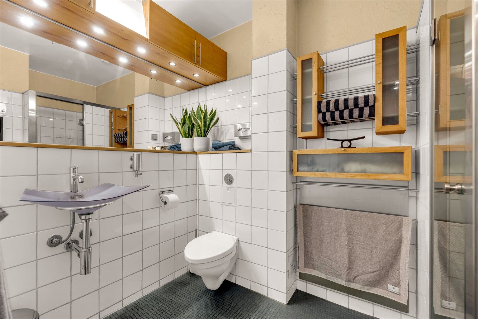 Badet er innredet med vegghengt servant med armatur, speil med overlys og praktisk baderomshylle under. Vegghengt skap ovenfor speilet samt over toalettet. 
