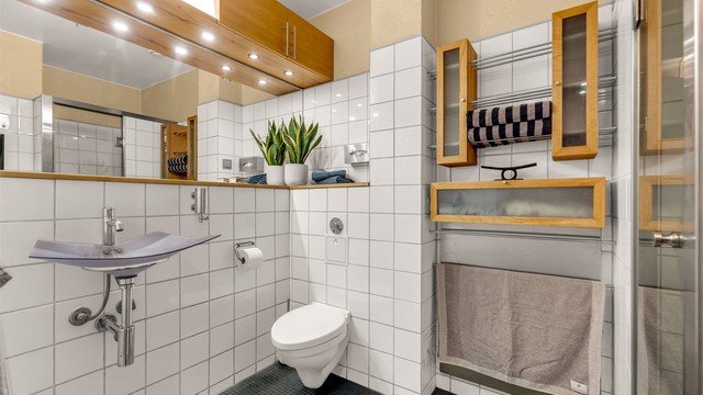 Badet er innredet med vegghengt servant med armatur, speil med overlys og praktisk baderomshylle under. Vegghengt skap ovenfor speilet samt over toalettet.