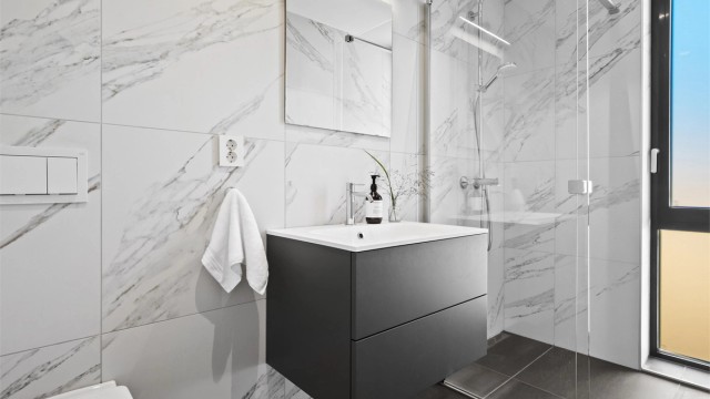 Badet har flislagte gulv og vegger, innredet med servant, vegghengt toalett og dusjhjørne.