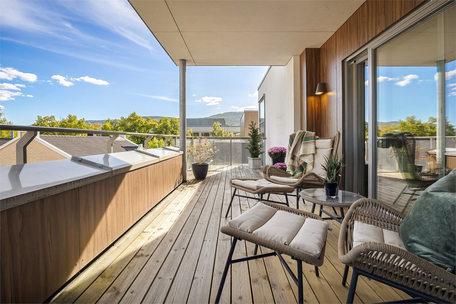Balkong med utvendig belysning, terrassebord på gulv og rekkverk av metall samt glass. 
