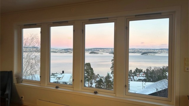 Selgers vinterfoto: Store vinduer i stuen gir flott utsikt mot Oslofjorden, med rosa lys og snødekte omgivelser som skaper en stemningsfull og unik atmosfære.