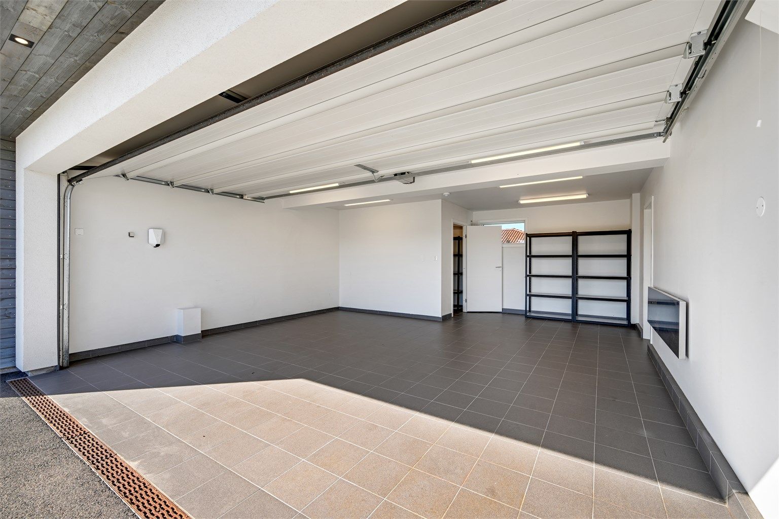 Integrert og isolert garasje med flislagt gulv og elbillader. Varmekabel er lagt i innkjørselen foran porten.