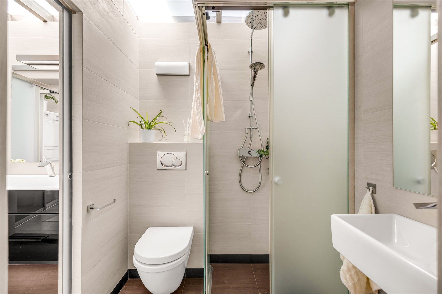Badet nærmest gangen er innredet med vegghengt toalett og dusjnisje i frostet glass, to-greps dusjarmatur på vegg og regnfallsdusj