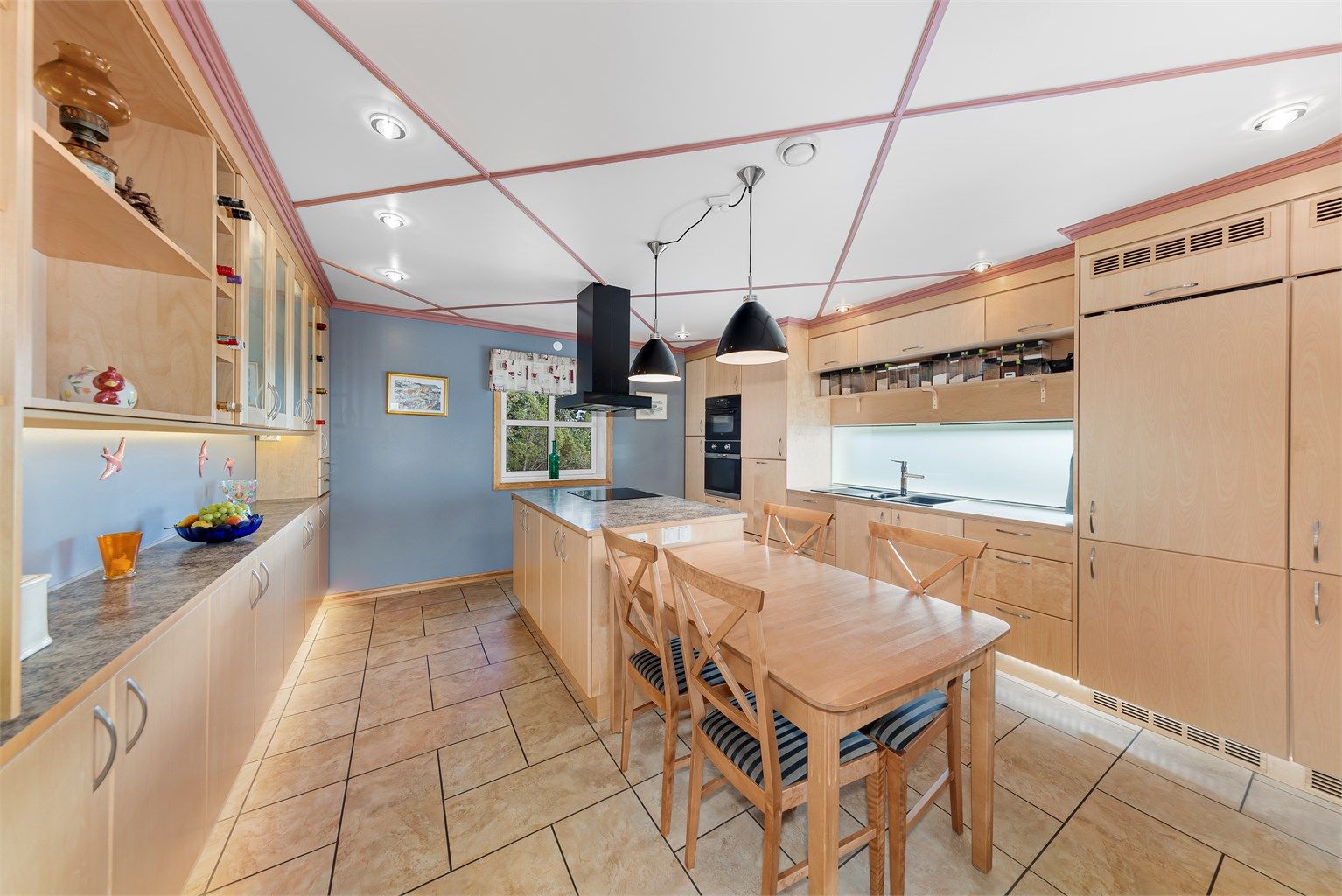 Kjøkkeninnredningen har integrerte hvitevarer og rikelig med skap- og benkeplass. Kjøkkenøya som er plassert midt i bildet har steketopp med ventilator over.