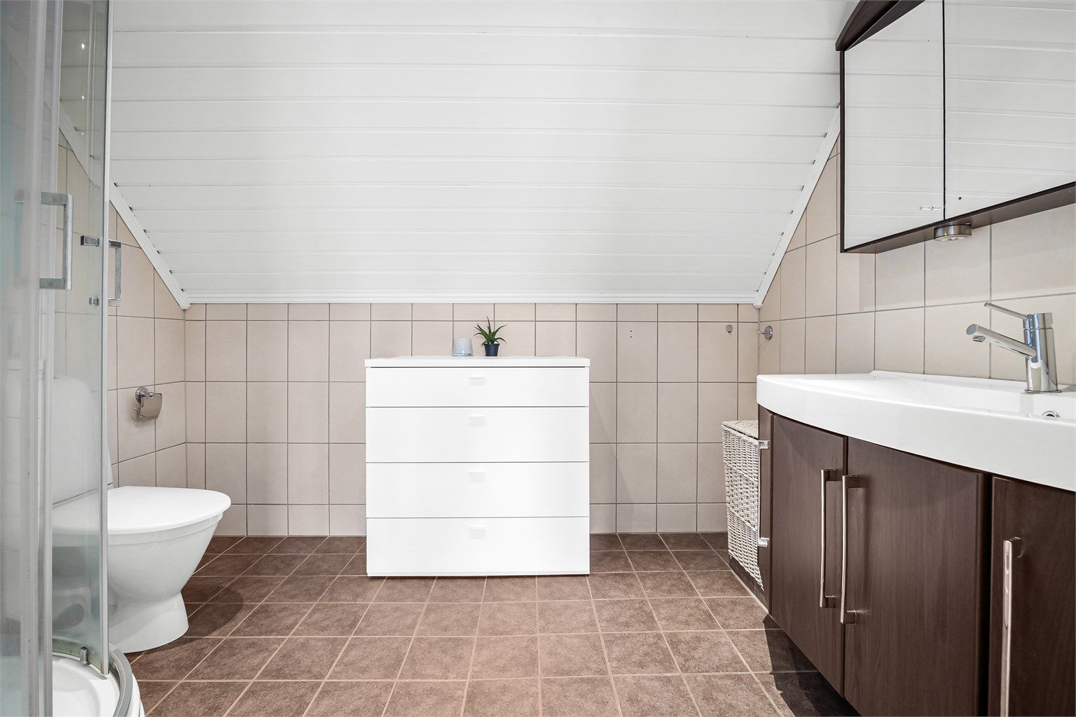 Badet er innredet med vegghengt servantinnredning og speilskap med overlys og stikkontakt på vegg over servant. Videre er badet utstyrt med dusjkabinett med skyvedører og gulvstående toalett. 