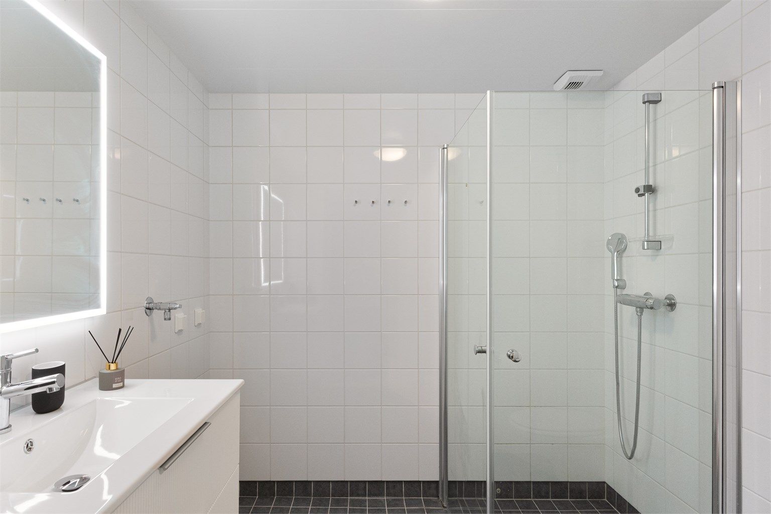 Lyst og flislagt baderom med behagelig gulvvarme og romslig dusjsone med innfellbare glassdører. Hvite veggfliser og mørkt gulv gir et rent og tidløst uttrykk.