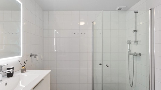 Lyst og flislagt baderom med behagelig gulvvarme og romslig dusjsone med innfellbare glassdører. Hvite veggfliser og mørkt gulv gir et rent og tidløst uttrykk.