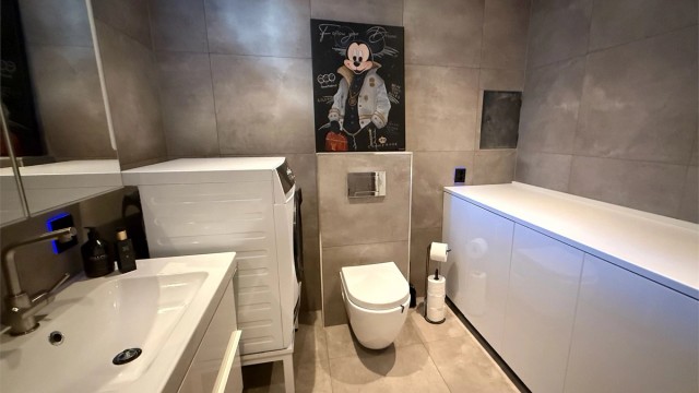 Badet er utstyrt med vegghengt toalett, servantinnredning og et nedsenket dusjhjørne med glassdører.