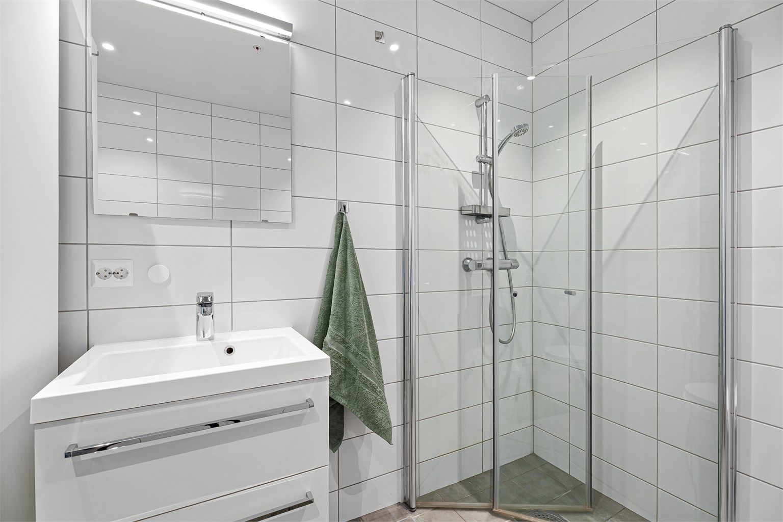 Badet har dusjnisje med innfellbare dører, vegghengt servant med skuffer samt speil med belysning. 