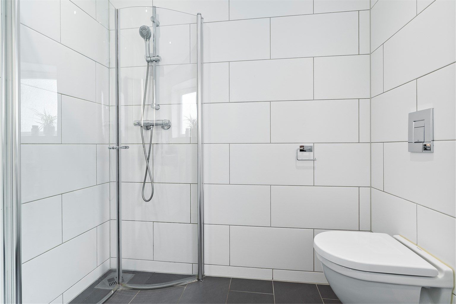 Dusjhjørnet har plassbesparende, foldbare dører og stilren armatur. Videre er det veggmontert toalett, opplegg for både vaskemaskin og tørketrommel, samt mekanisk avtrekk fra badet