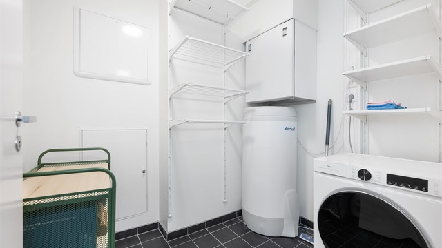 Praktisk separat vaskerom med flislagt gulv, opplegg for vaskemaskin, varmtvannsbereder og gode oppbevaringshyller.