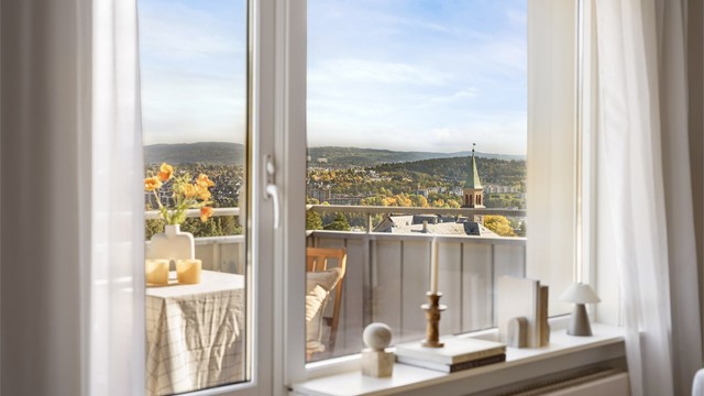 Kirketårnet stikker opp mellom trærne, og i horisonten ligger det grønne åslandskapet. Den samme utsikten får du fra både sofaen og spisebordet, og høyt oppe i tolvte etasje er den din hele året.
