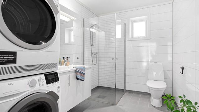 Tidløst og innholdsrikt bad med fliser på gulv og gulvvarme samt baderomsplater på vegg. Videre er det dusnisje med innfellbare dører og toalett.