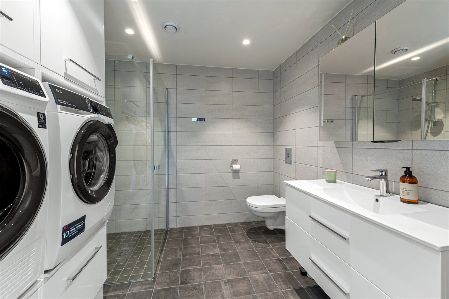 Bad 2/vaskerom med tilkomst fra gang. Innredning med servant, speilskap, vegghengt wc og dusjhjørne med vegghengt armatur. Innredning med opplegg for vaskemaskin og tørketrommel