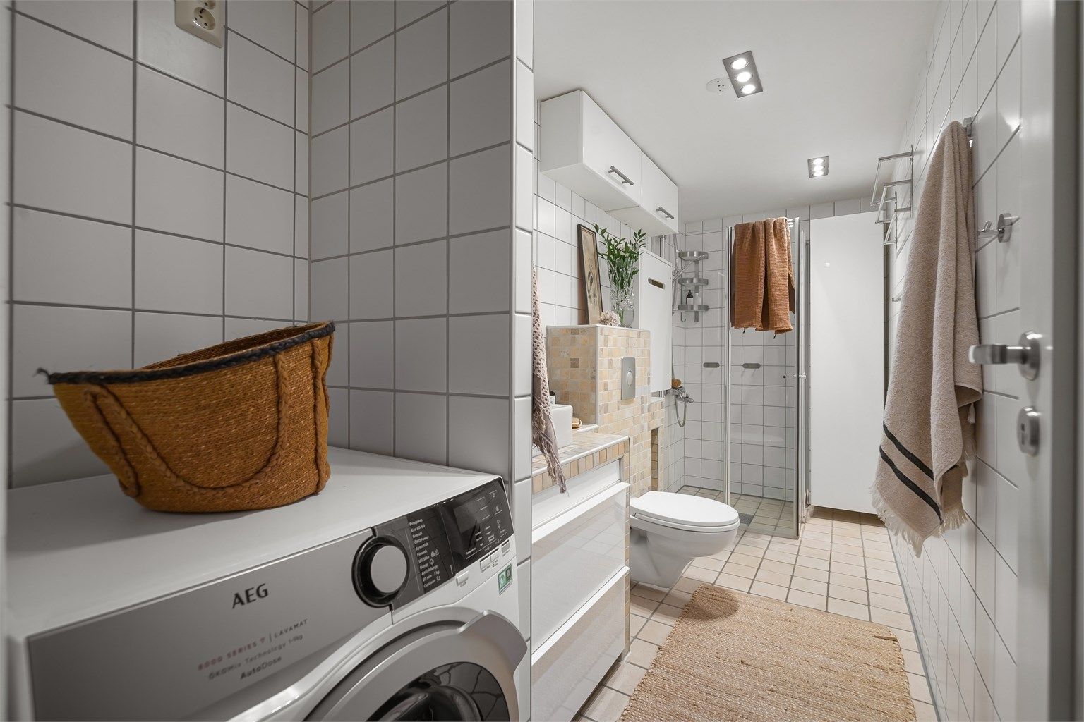 Innbydende og meget romslig bad med hvite fliser på vegg, beigemelerte fliser på gulv med varmekabler og downlights i himling