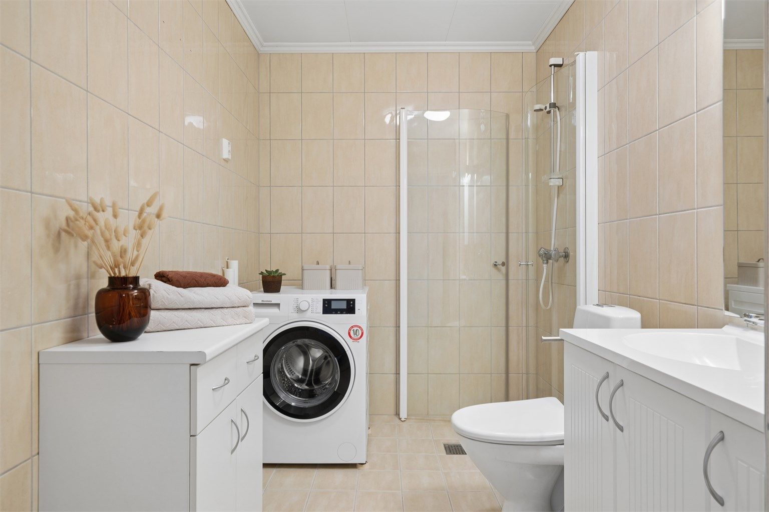 Badet har flislagte gulv og vegger, gulvvarme, dusjhjørne, vegghengt servantinnredning og toalett.