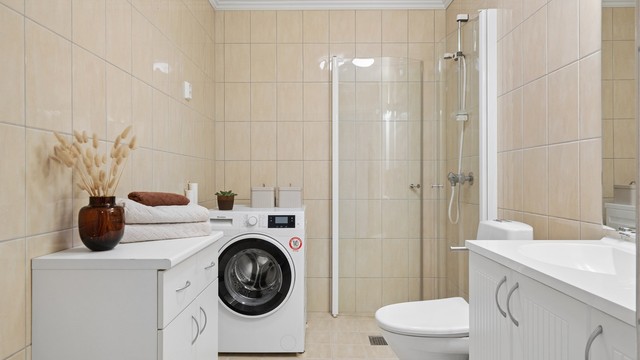 Badet har flislagte gulv og vegger, gulvvarme, dusjhjørne, vegghengt servantinnredning og toalett.