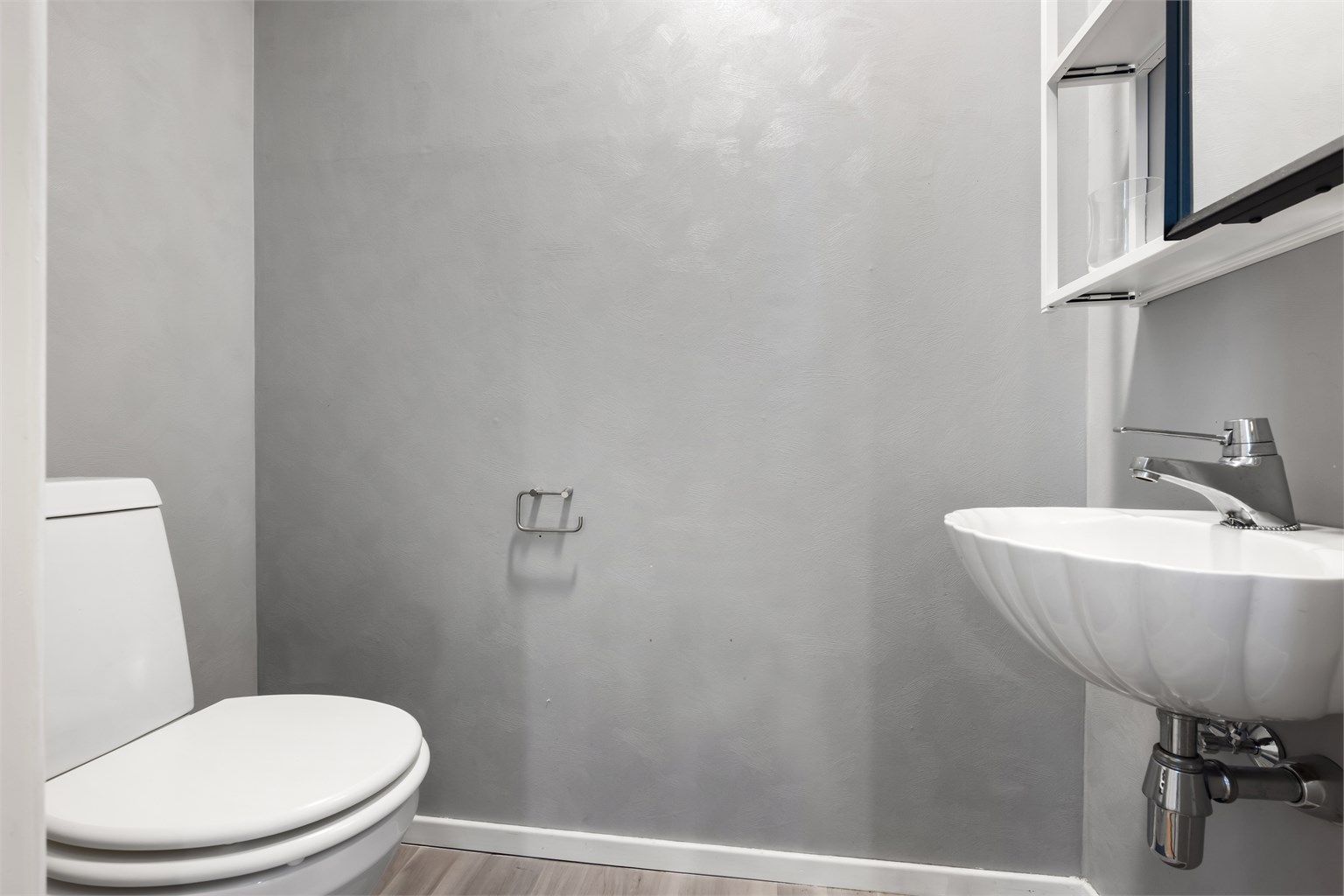 Toalettrom fra byggeår og vegghengt servant med ett-greps armatur. Det er speilskap på vegg over servant, gulvstående toalettet fra omkring 2022 og avtrekksventil plassert i himling.