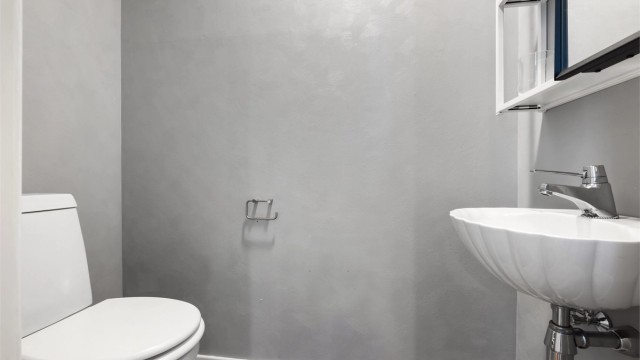 Toalettrom fra byggeår og vegghengt servant med ett-greps armatur. Det er speilskap på vegg over servant, gulvstående toalettet fra omkring 2022 og avtrekksventil plassert i himling.