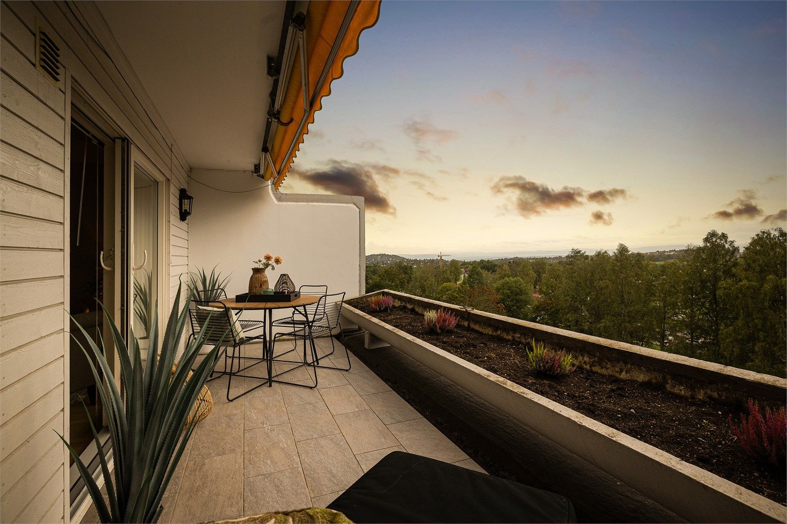 Vestvendt balkong på 11 kvm, belagt med fliser og utstyrt med utebelysning, markise og rekkverk med integrert blomsterkasse.