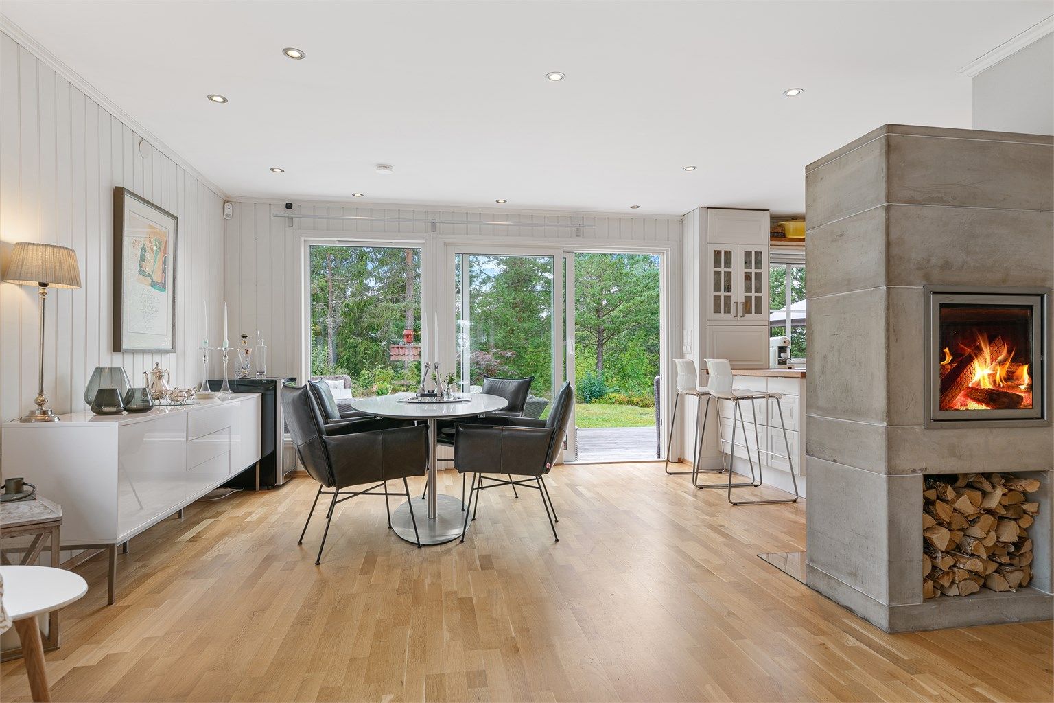 Lyst og sosialt allrom med store vindusflater, klebersteinspeis og åpen løsning mot kjøkkenet. Skyvedør gir direkte adkomst til solrik terrasse og hage