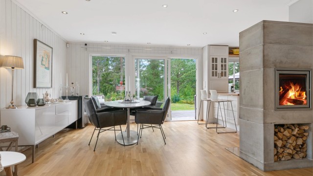 Lyst og sosialt allrom med store vindusflater, klebersteinspeis og åpen løsning mot kjøkkenet. Skyvedør gir direkte adkomst til solrik terrasse og hage