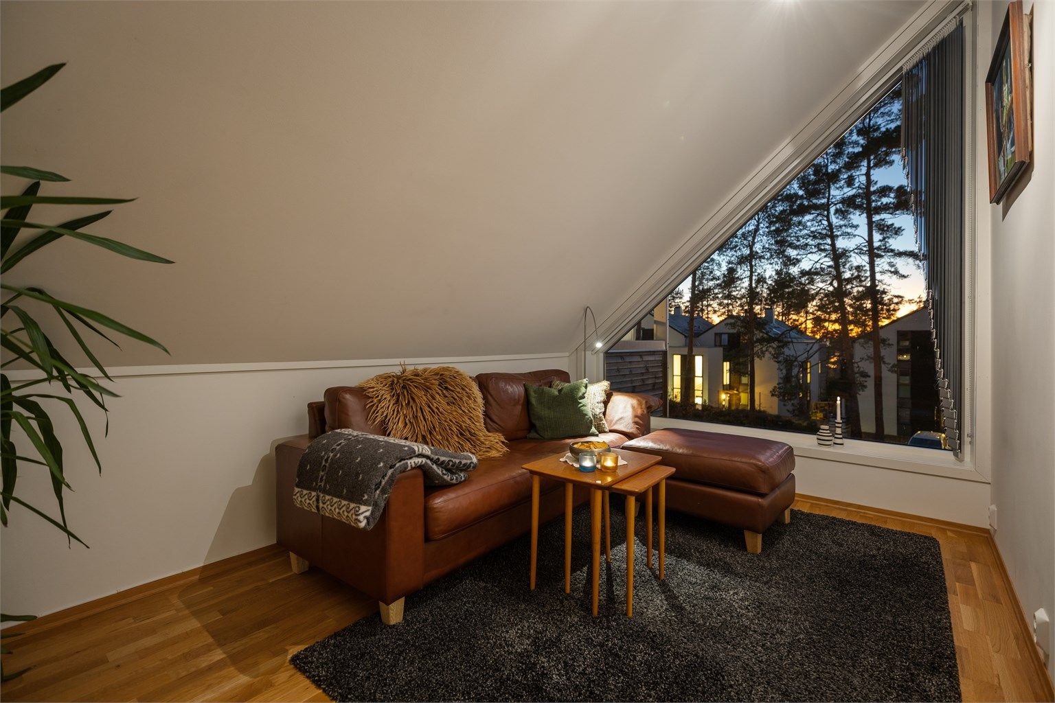 Lun og innbydende loftstue med flere bruksmuligheter, i dag innredet som TV-stue – en skjermet sone for avslapning og underholdning. Det særpregede vinduspartiet slipper inn naturlig lys og gir en stemningsfull utsikt mot kveldshimmelen.