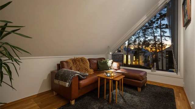 Lun og innbydende loftstue med flere bruksmuligheter, i dag innredet som TV-stue – en skjermet sone for avslapning og underholdning. Det særpregede vinduspartiet slipper inn naturlig lys og gir en stemningsfull utsikt mot kveldshimmelen.