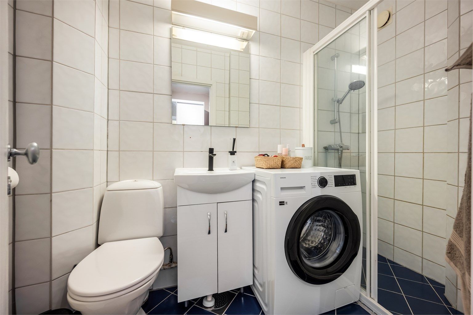 Flislagt bad med dusjhjørne med skyvedør av glass og opplegg for vaskemaskin. Varmekabler er defekte.