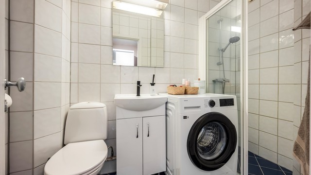 Flislagt bad med dusjhjørne med skyvedør av glass og opplegg for vaskemaskin. Varmekabler er defekte.