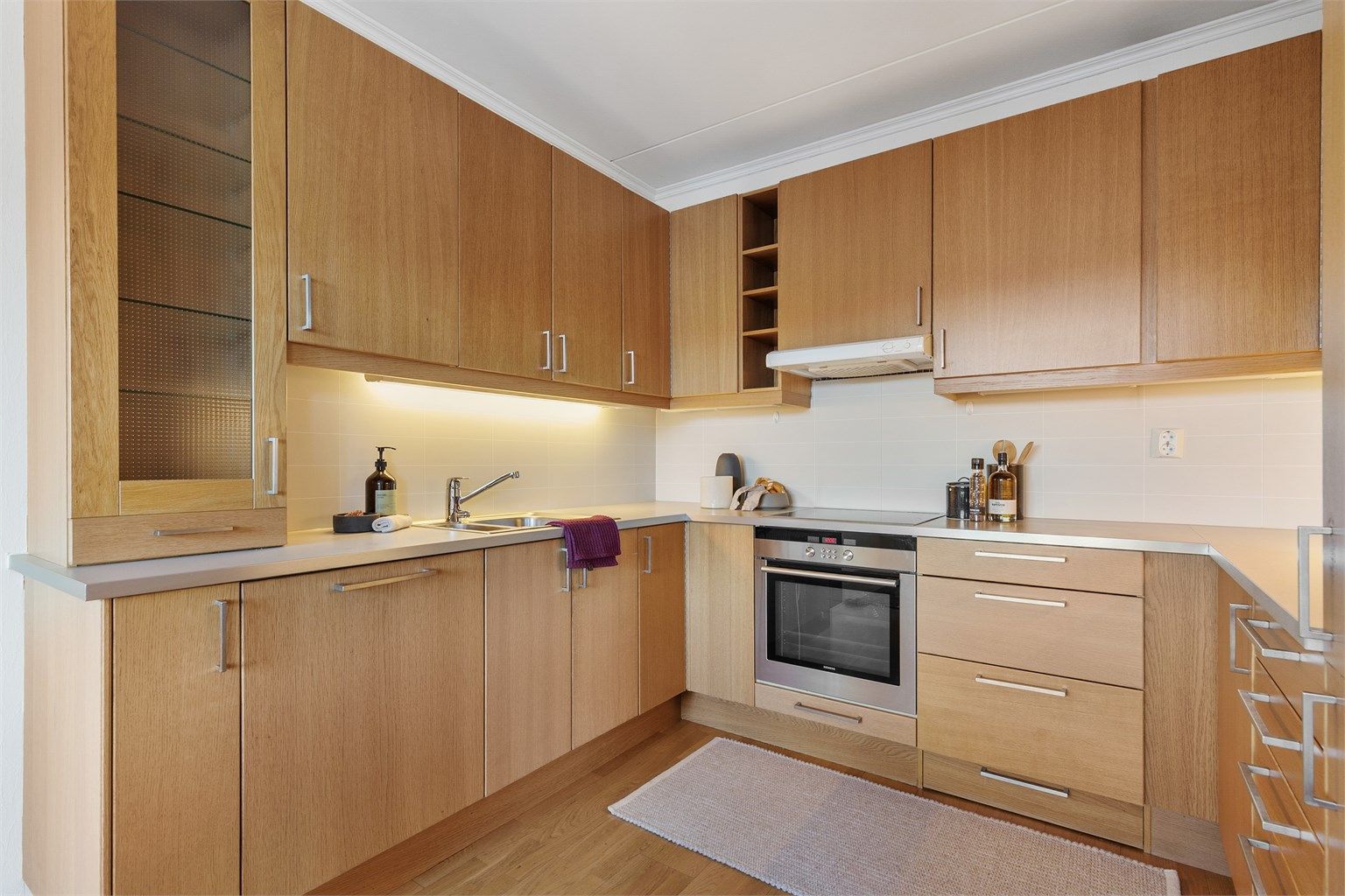 Åpen kjøkkenløsning. Innredning fra byggeår. Glatte fronter. Benkeplate med laminert overflate. Kjøkkenvask med armatur. Stikkontakt over kjøkkenbenk. Integrert stekeovn, platetopp, oppvaskmaskin og kjøleskap med fryser.