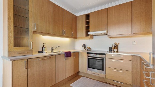 Åpen kjøkkenløsning. Innredning fra byggeår. Glatte fronter. Benkeplate med laminert overflate. Kjøkkenvask med armatur. Stikkontakt over kjøkkenbenk. Integrert stekeovn, platetopp, oppvaskmaskin og kjøleskap med fryser.