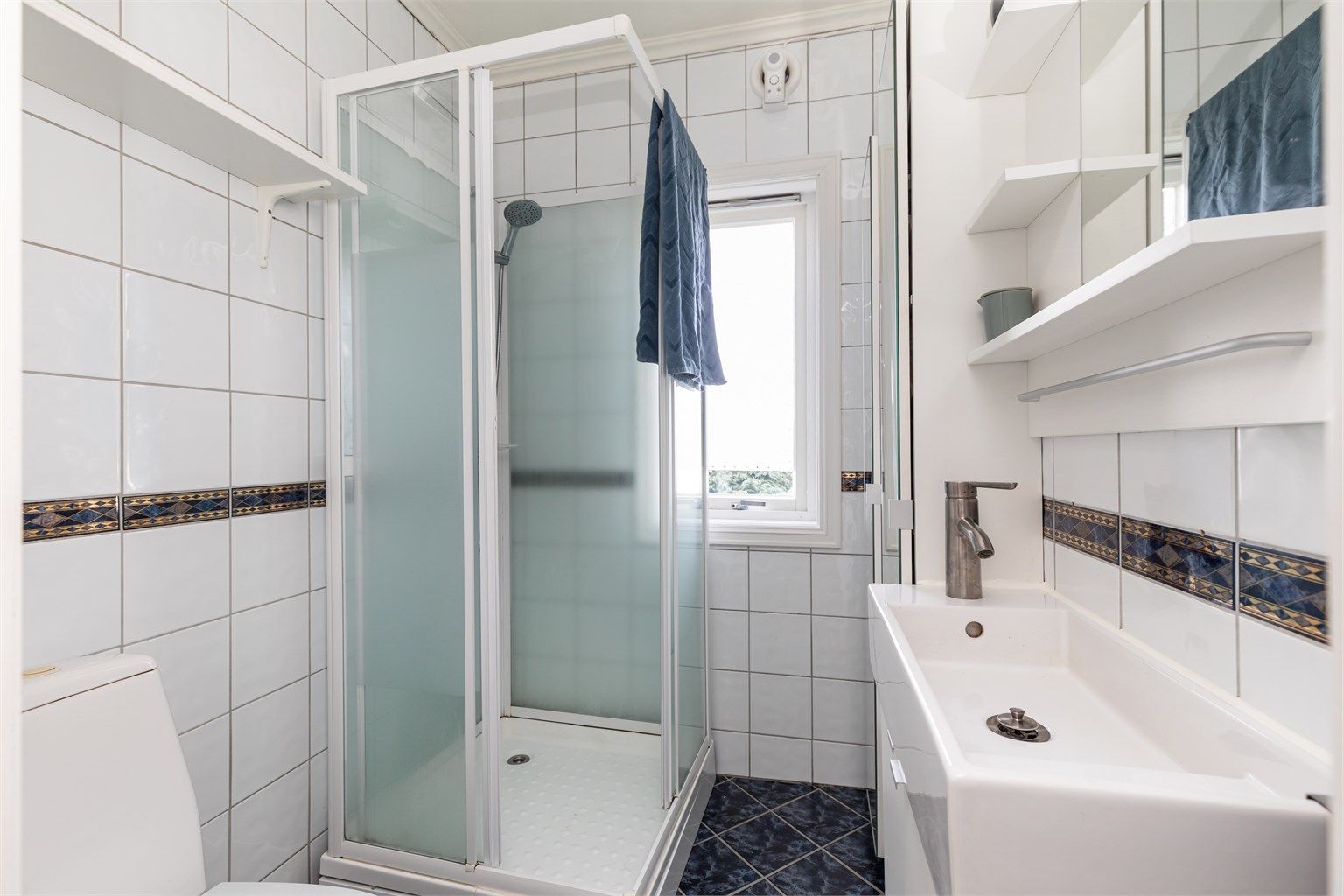 Badet har flislagte vegger og gulv, utstyrt med dusjkabinett og vindu for naturlig dagslys.