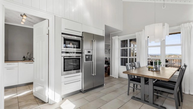 Åpen kjøkkenløsning. Innredning fra byggeår. Profilerte fronter. Benkeplate av tre og stein. Nedfelt kjøkkenvask med armatur. Stikkontakter over kjøkkenbenk. Frittstående kjøleskap med fryser.