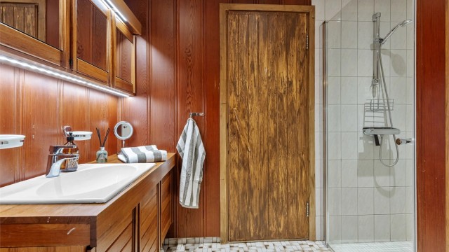 Baderom fra byggeår. Flislagt gulv med gulvvarme. Panel på vegger. Flislagte vegger i dusjsone. Panelbord med downlights i himling. Vegghengt servantinnredning. Benkeplate av tre. Nedfelt servant med armatur. Speilskap med belysning. Gulvstående toalett.