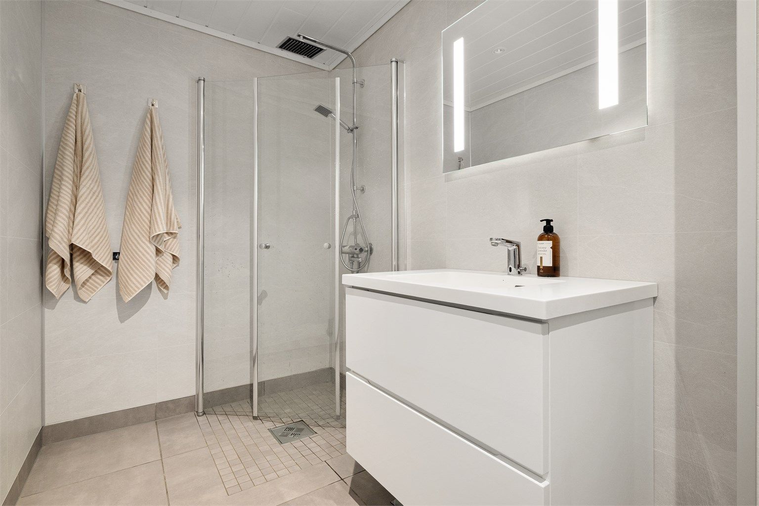 Badet er utstyrt med pen vegghengt servantinnredning, dusjhjørne med glassdører og vegghengt toalett.