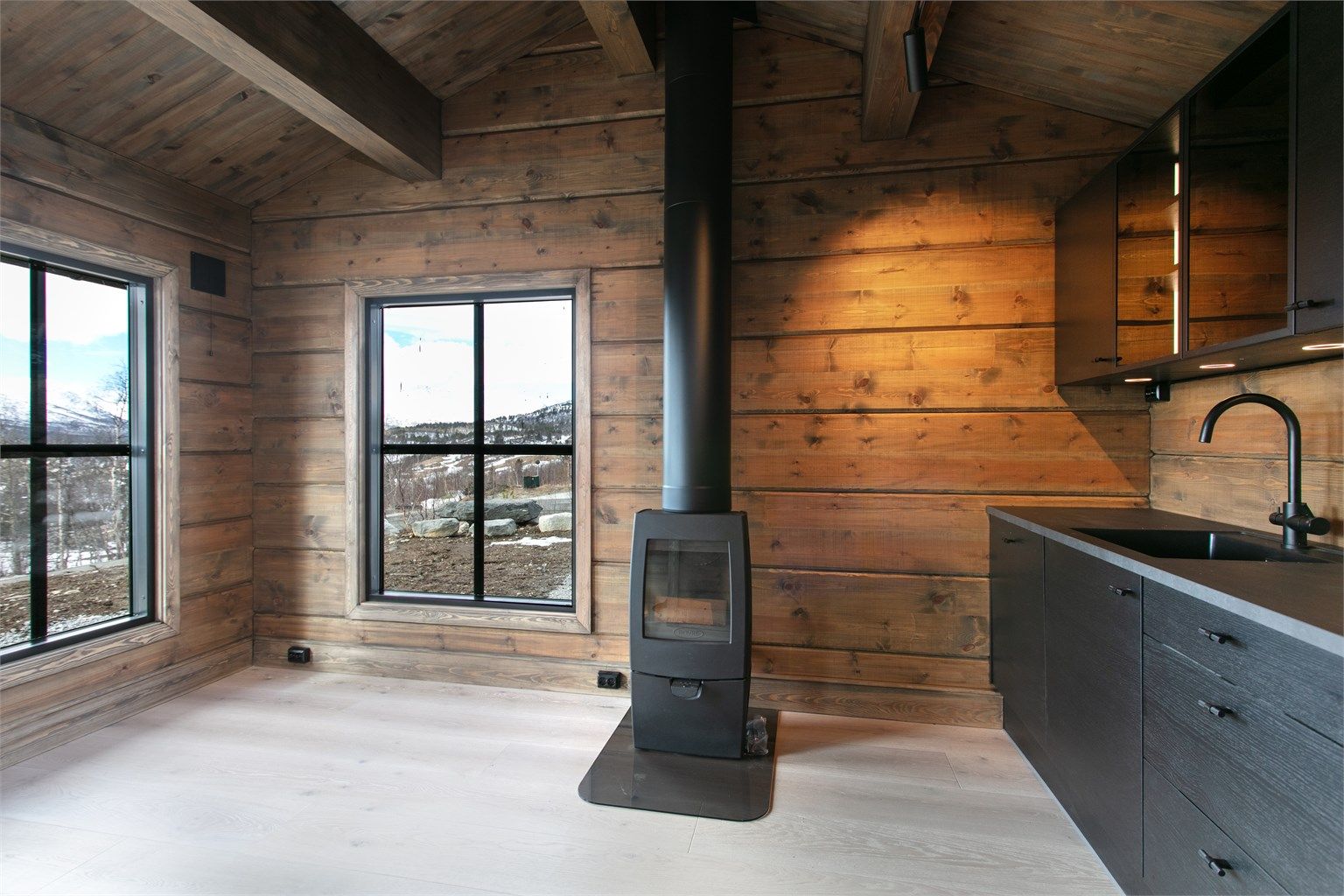 Kombinasjonen av det mørke kjøkkenet, tømmerveggene og peisovnen skaper en moderne og samtidig tradisjonell fjellhyttefølelse.