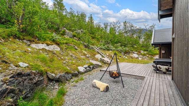 Skjermet uteplass på baksiden av hytta med gruset parti og bålplass mot naturtomten.