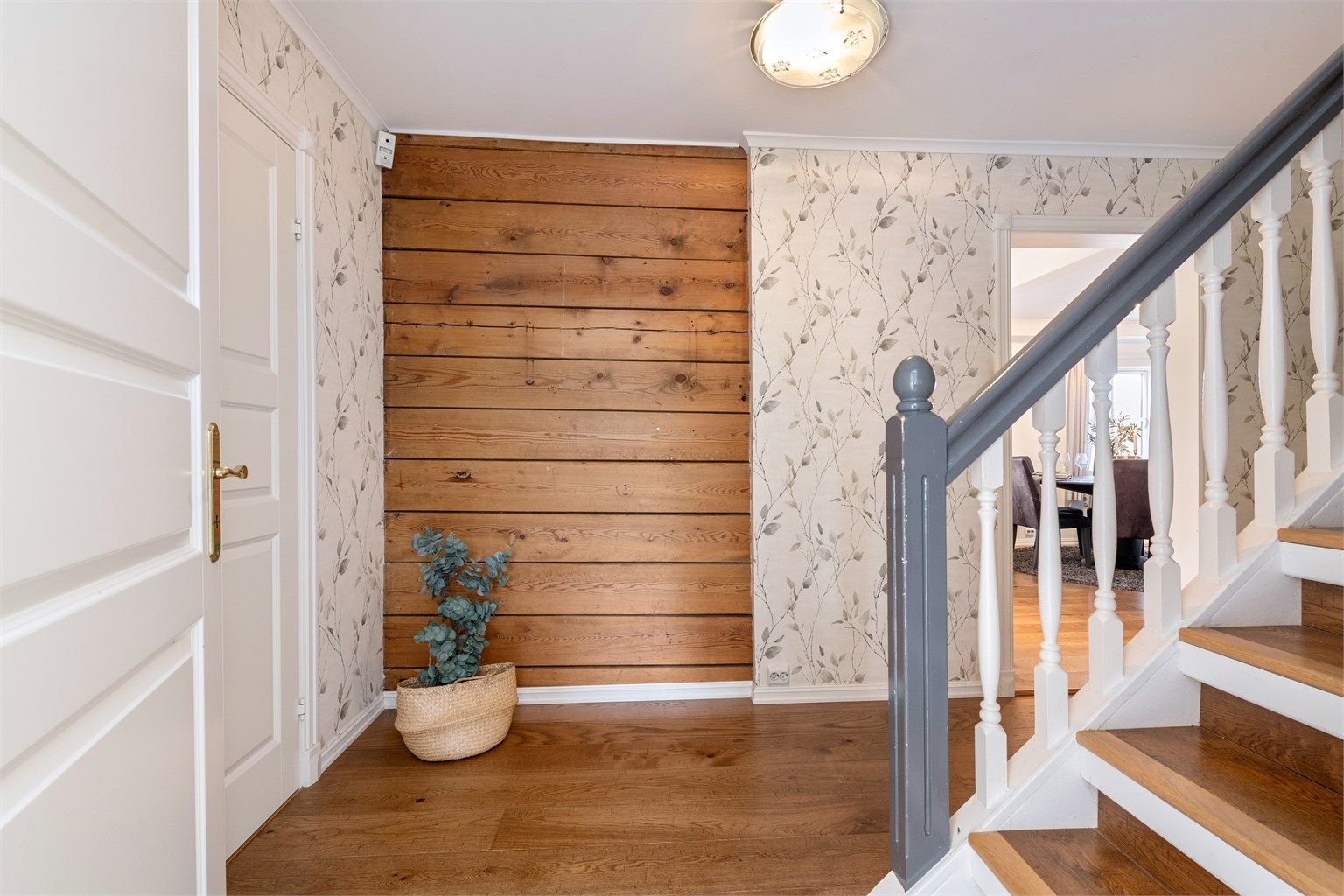 Gang/trapperom med parkett på gulv, vegger har tapet og noe synlige tømmervegger. Malt himling. Innvendig trapp i tre mellom hovedtasje og loft
