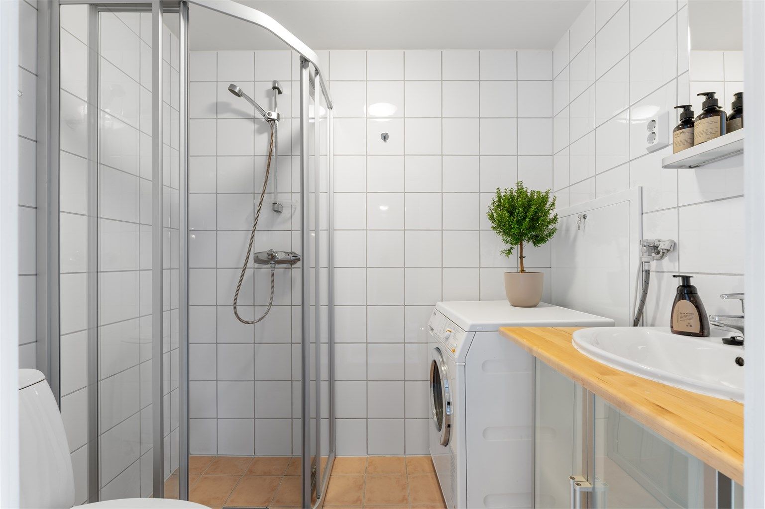 Baderom med flislagte overflater, gulvvarme og opplegg for vaskemaskin. Servantinnredning med overliggende speil og tilhørende hylle.
