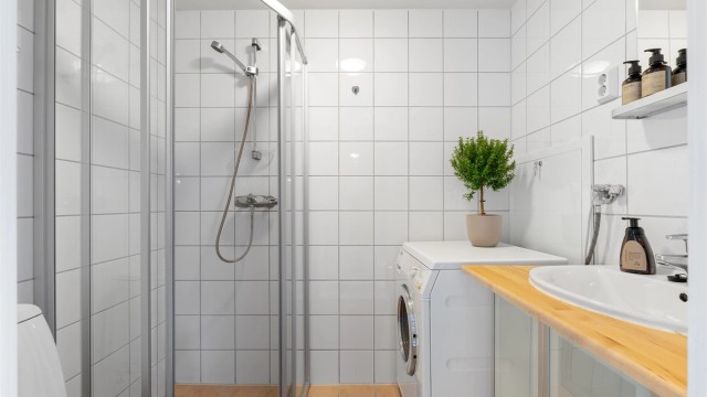 Baderom med flislagte overflater, gulvvarme og opplegg for vaskemaskin. Servantinnredning med overliggende speil og tilhørende hylle.