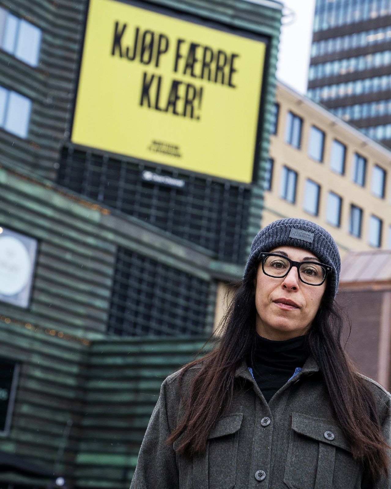 Woman standing in front of advertising sign saying "Buy less clothes!"