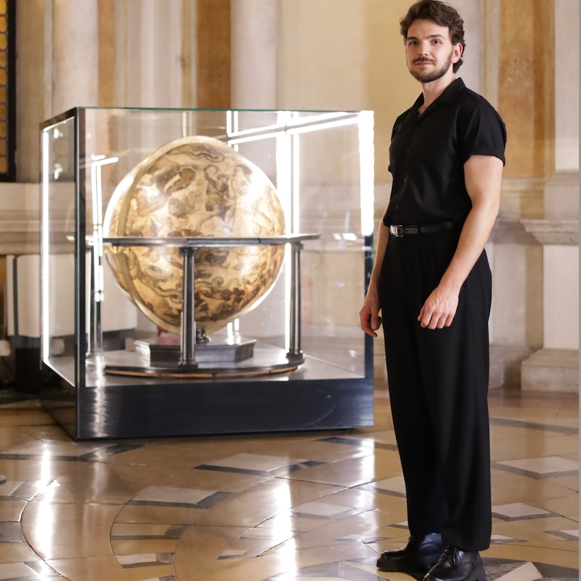 Leonardo Frigo stands in the Marciana Library beside a celestial globe by Vincenzo Coronelli, displayed in a glass case within the ornate marble hall.