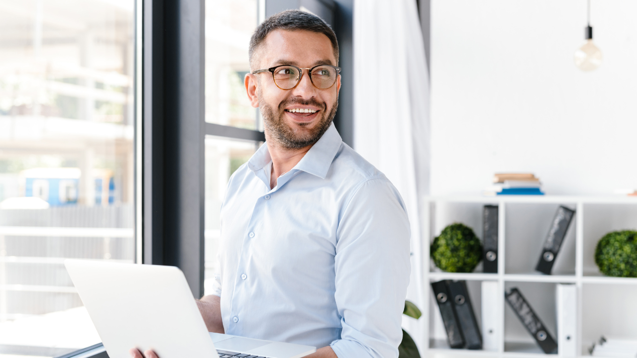 Man smiling while working on a laptop in a bright office.