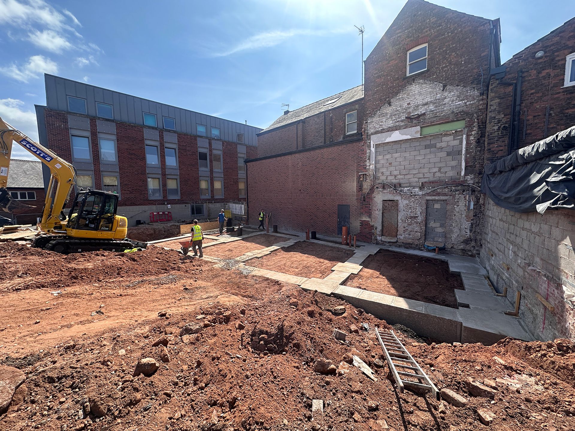 The cleared site with strip foundations poured and an excavator levelling ground.